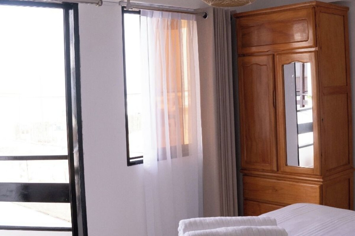 Ein Hotelzimmer mit einem Bett, einem Holzschrank, einem großen Fenster mit Vorhängen und einer gewobenen Deckenleuchte.