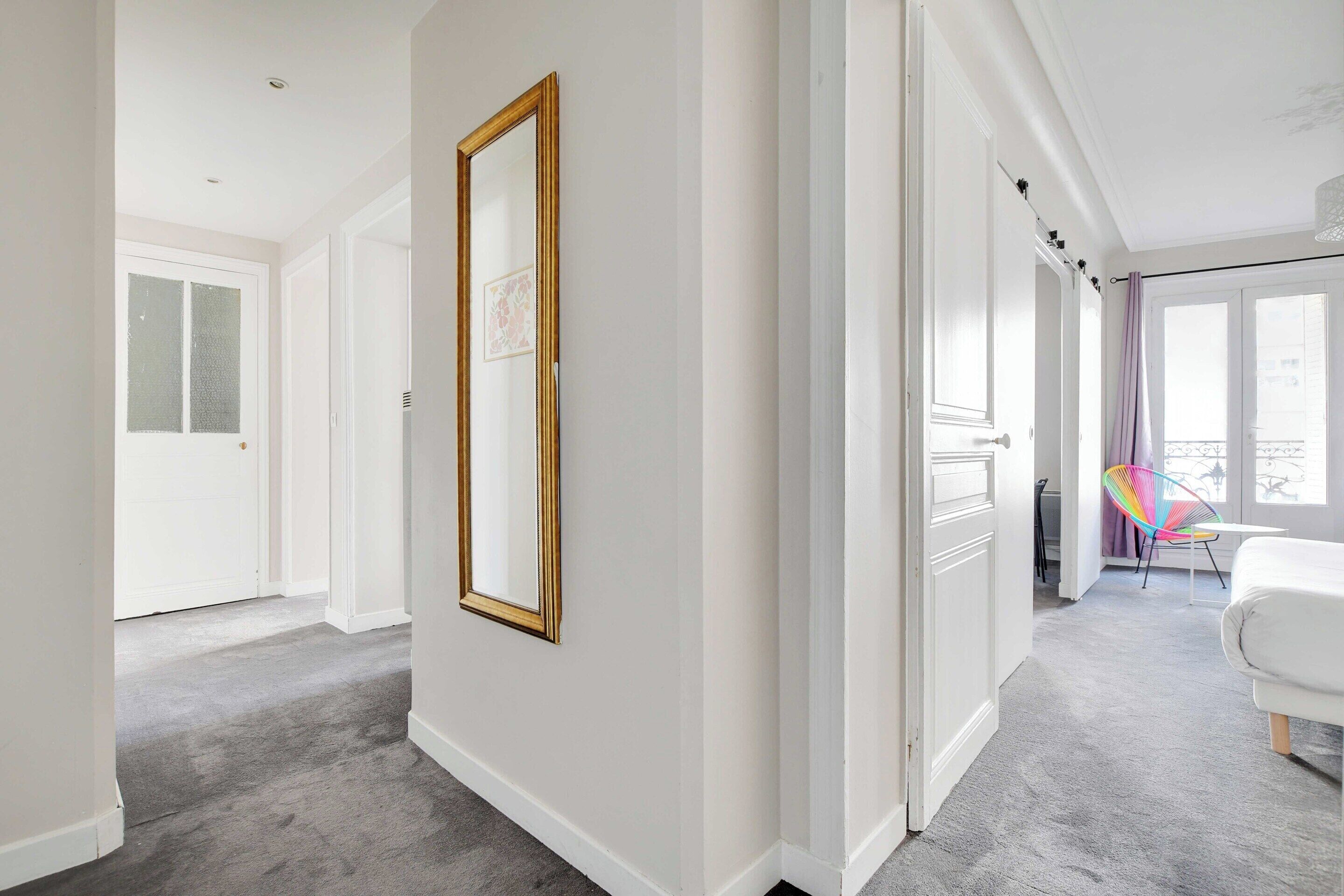 A hallway with a large mirror, a white door, and a view into a room with a bed and a chair.