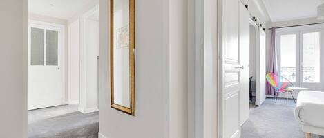 A hallway with a large mirror, a white door, and a view into a room with a bed and a chair.