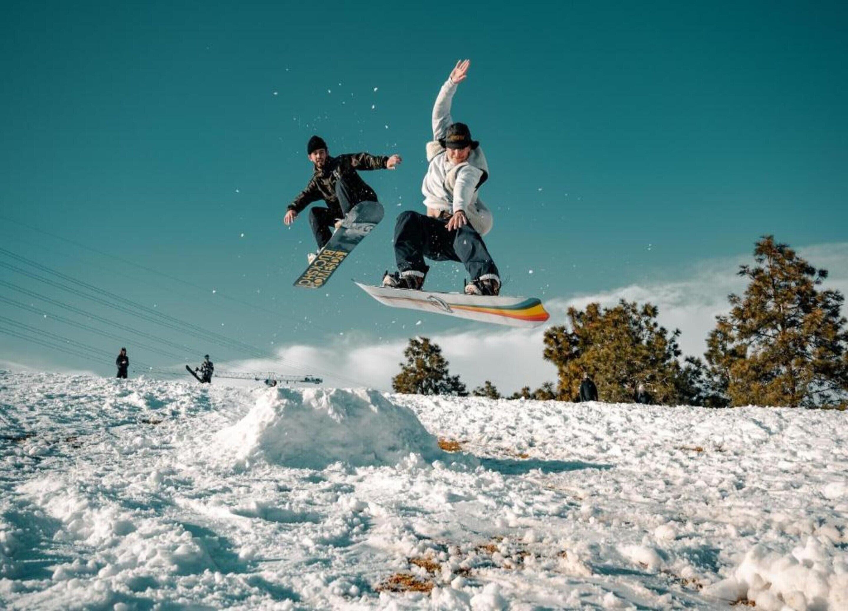 Un snowboarder en plein saut, au-dessus d’une pente enneigée, avec des arbres en arrière-plan.