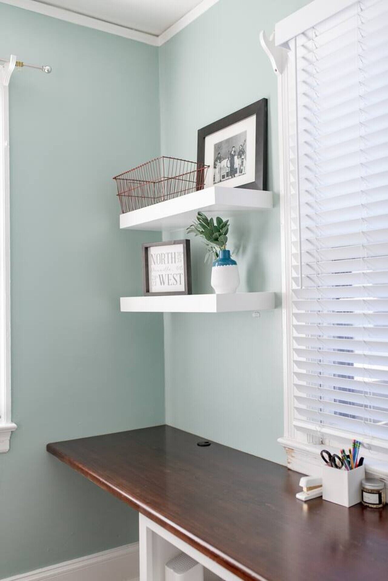 A corner desk with a dark wood top, white shelves, a framed picture, a small plant, and a window with white blinds.