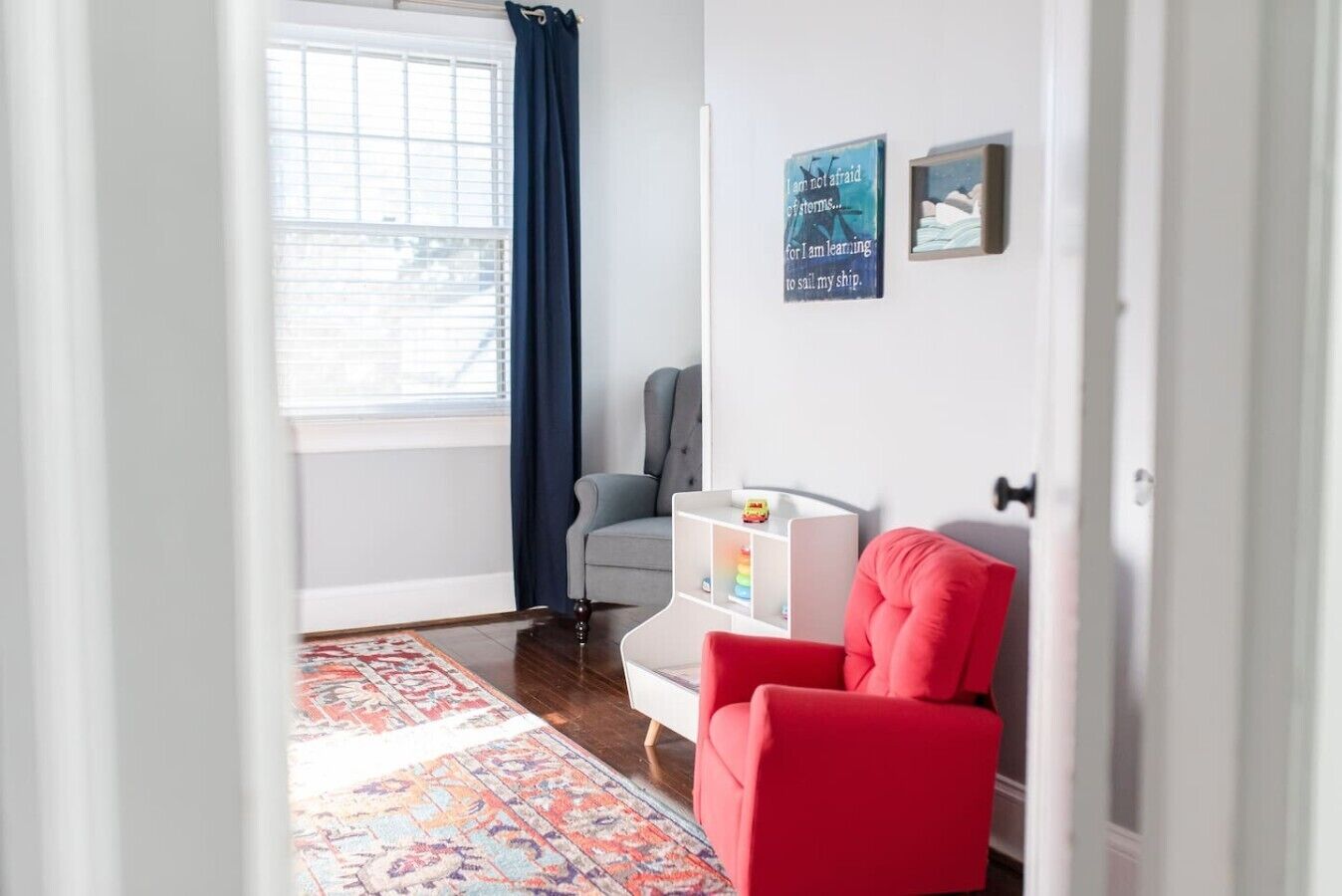 Ein modernes Wohnzimmer mit einem grauen Sofa, einem roten Sessel, einem weißen Bücherregal und einem blauen Vorhang.