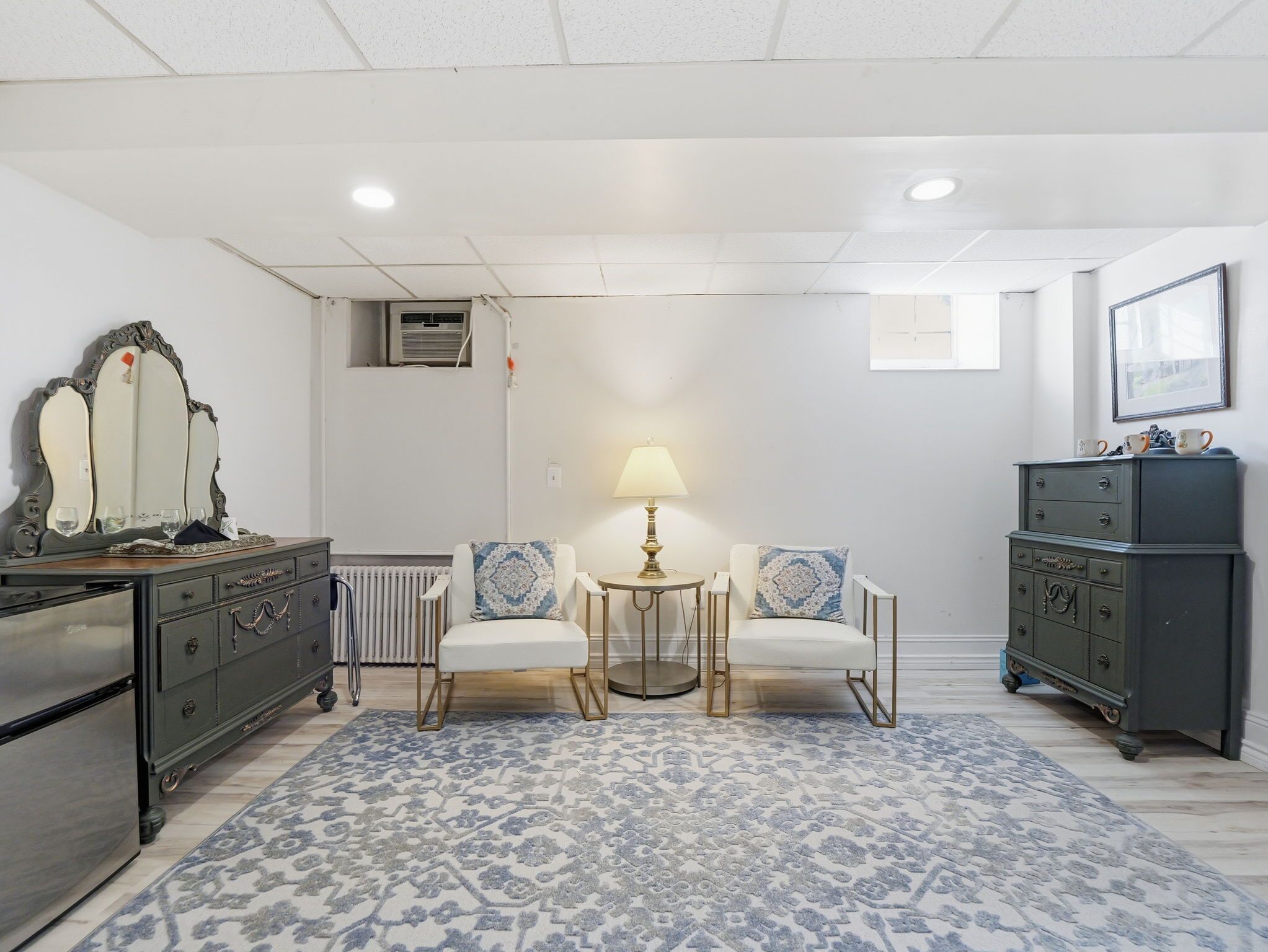 Una habitación con techo blanco, un radiador, un espejo, una mesa con lámpara, dos sillas, un tapete estampado y un aparador de color oscuro.