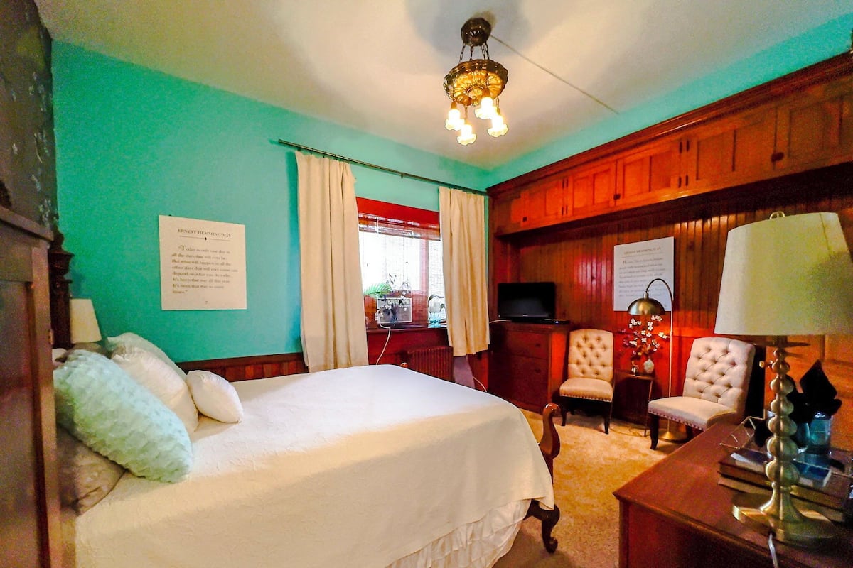 A hotel room with a bed, two armchairs, a desk with a lamp, a television, and a chandelier.