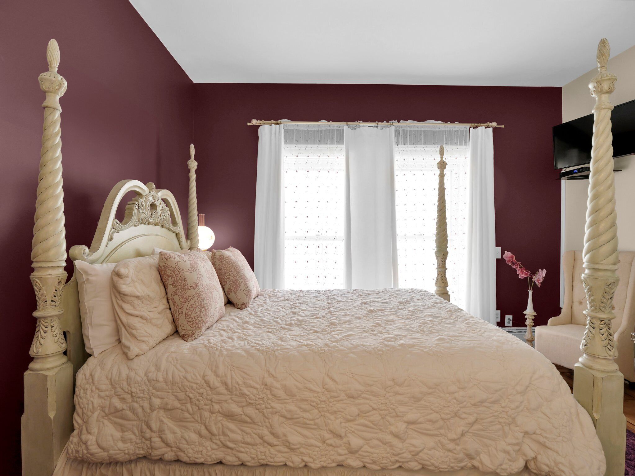Une chambre à coucher avec un lit à baldaquin, un téléviseur fixé au mur et une fenêtre ornée de rideaux légers.