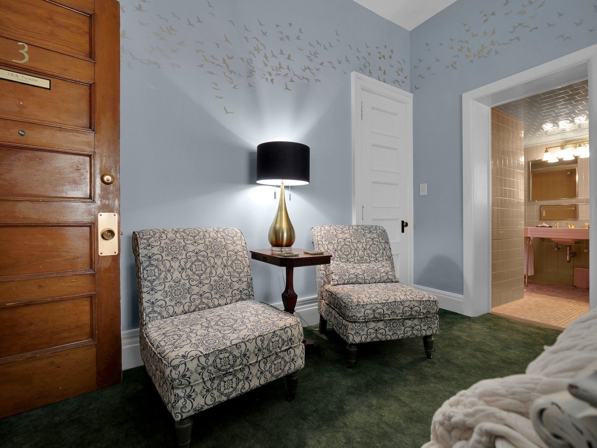 A room with two patterned armchairs, a small table with a lamp, a wooden door with a brass plate, and a bathroom visible through an open doorway.