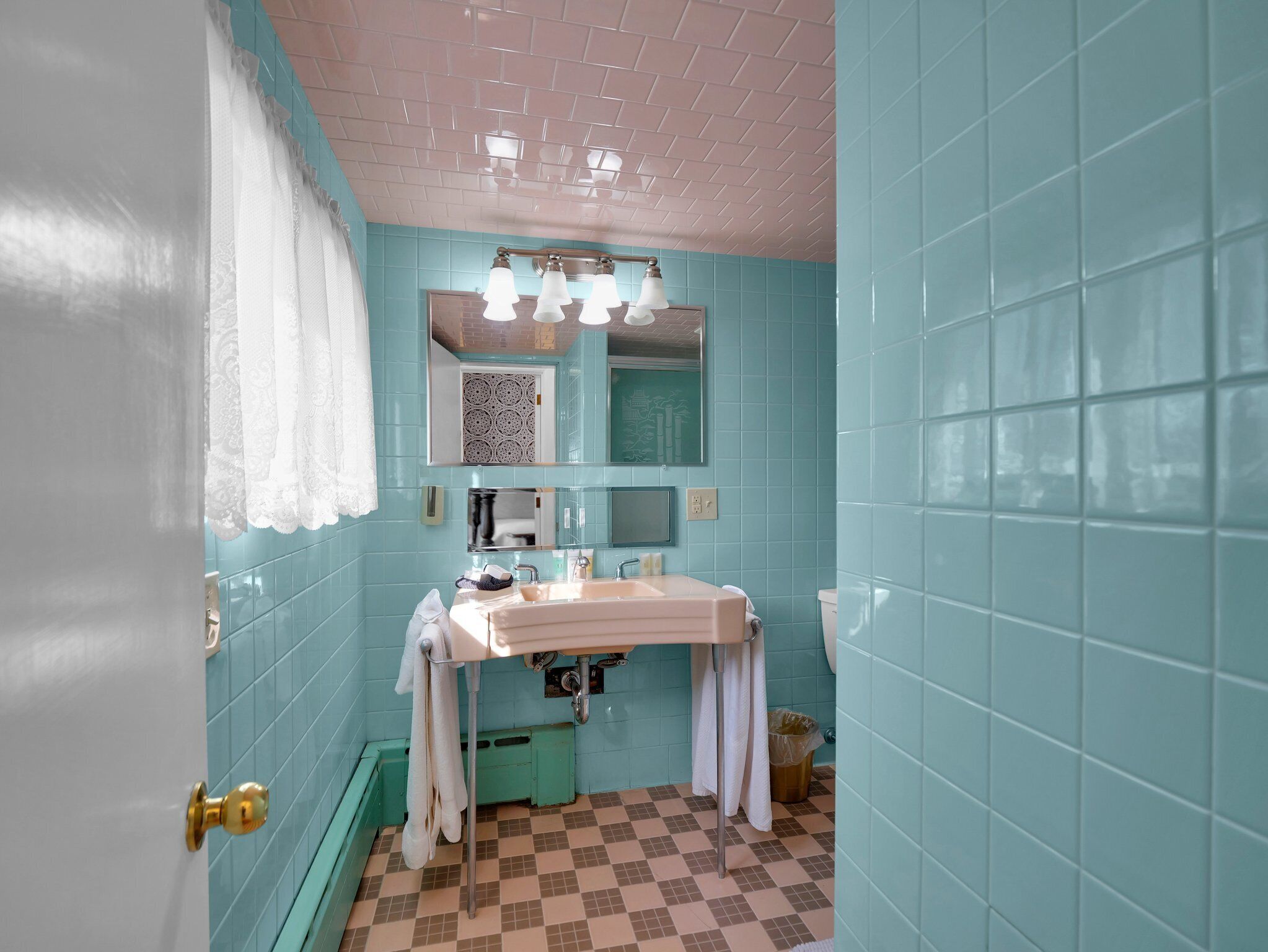 Baño con paredes verde azulado, un lavamanos blanco y un espejo encima. Se ve una ventana con cortina blanca y un cubo de basura.