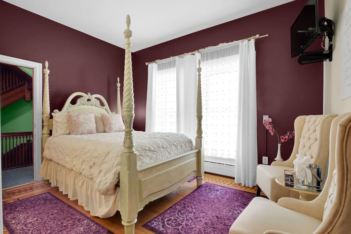 A bedroom with a four-poster bed, a sitting area with a chair, and a television mounted on the wall.