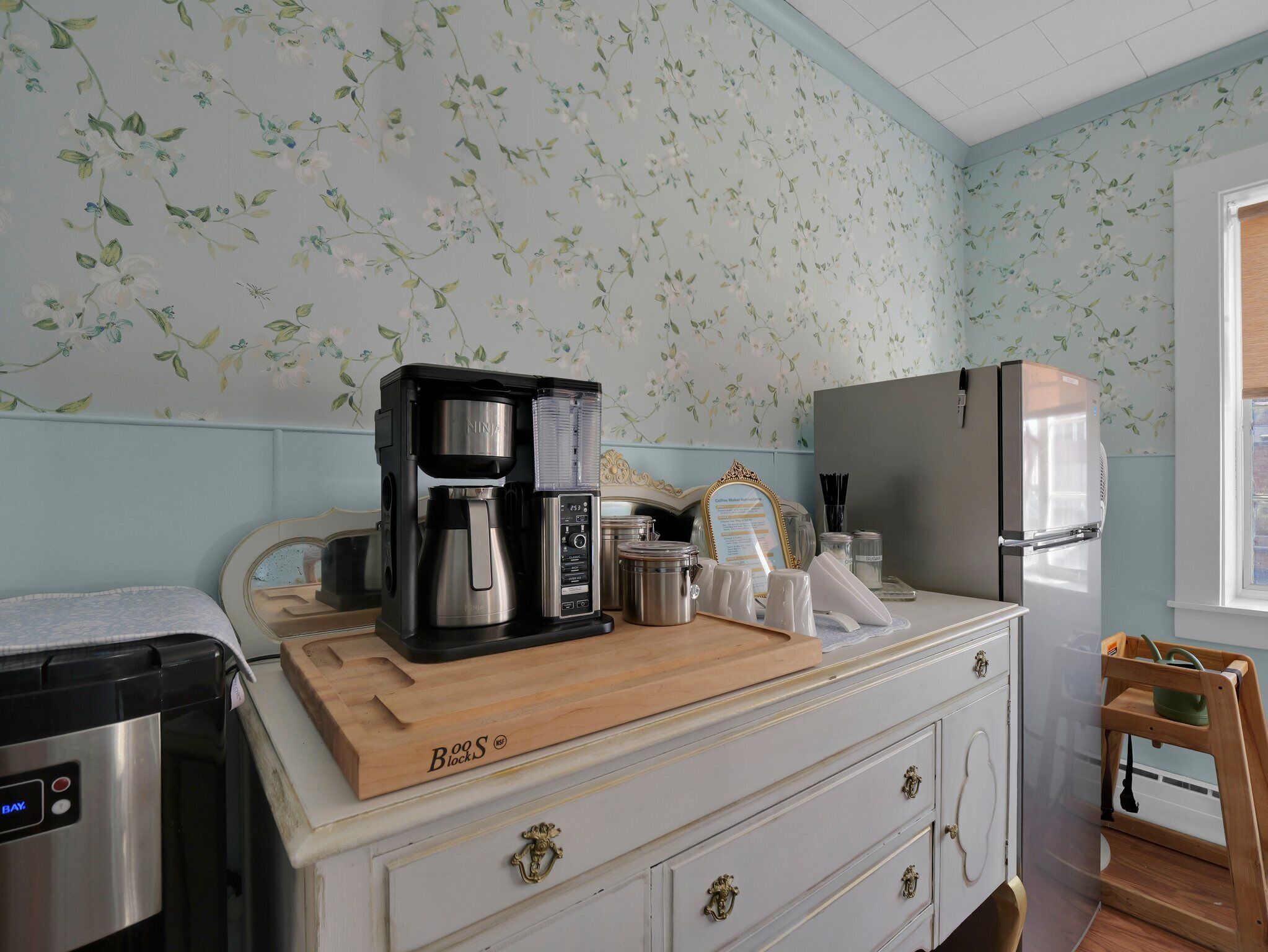 A kitchen with a coffee maker, refrigerator, and wooden cutting board on the counter.