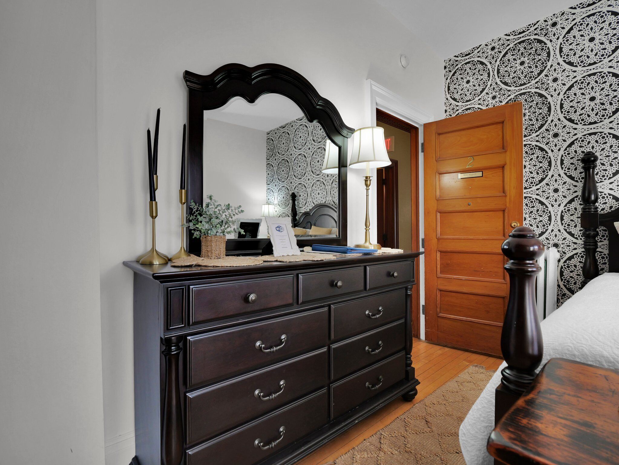 A bedroom with a dark wooden dresser, a large mirror, a bedside table with a lamp, and a patterned wallpaper.
