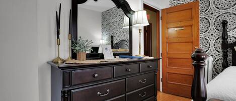 A bedroom with a dark wooden dresser, a large mirror, a bedside table with a lamp, and a patterned wallpaper.