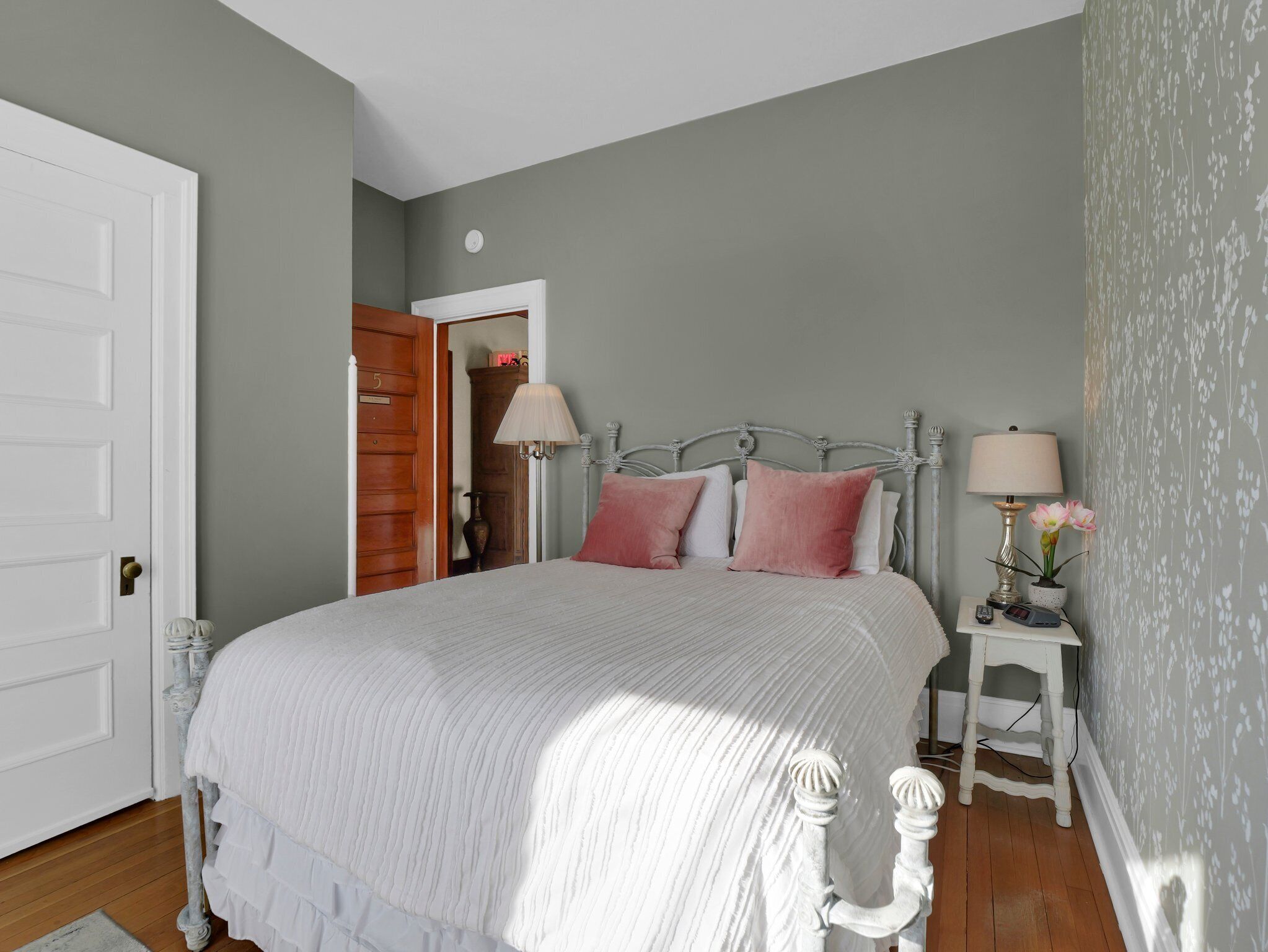 A bedroom with a large bed, two bedside tables, a nightstand, a door, and a wall with a floral pattern.