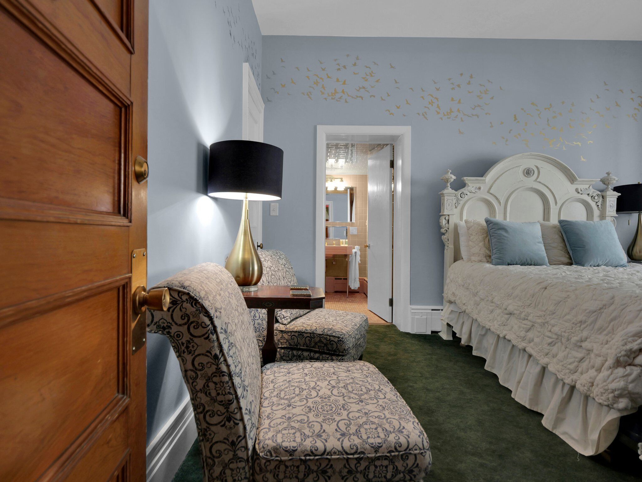 A bedroom with a wooden door, a patterned armchair, a bedside table with a lamp, a bed with a white headboard, and a bathroom visible through an open door.