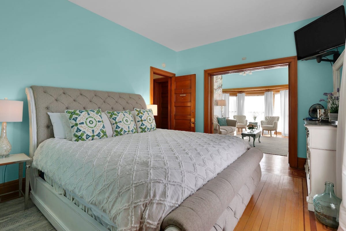 A bedroom with a large bed, a wooden door, and a view of a dining area.