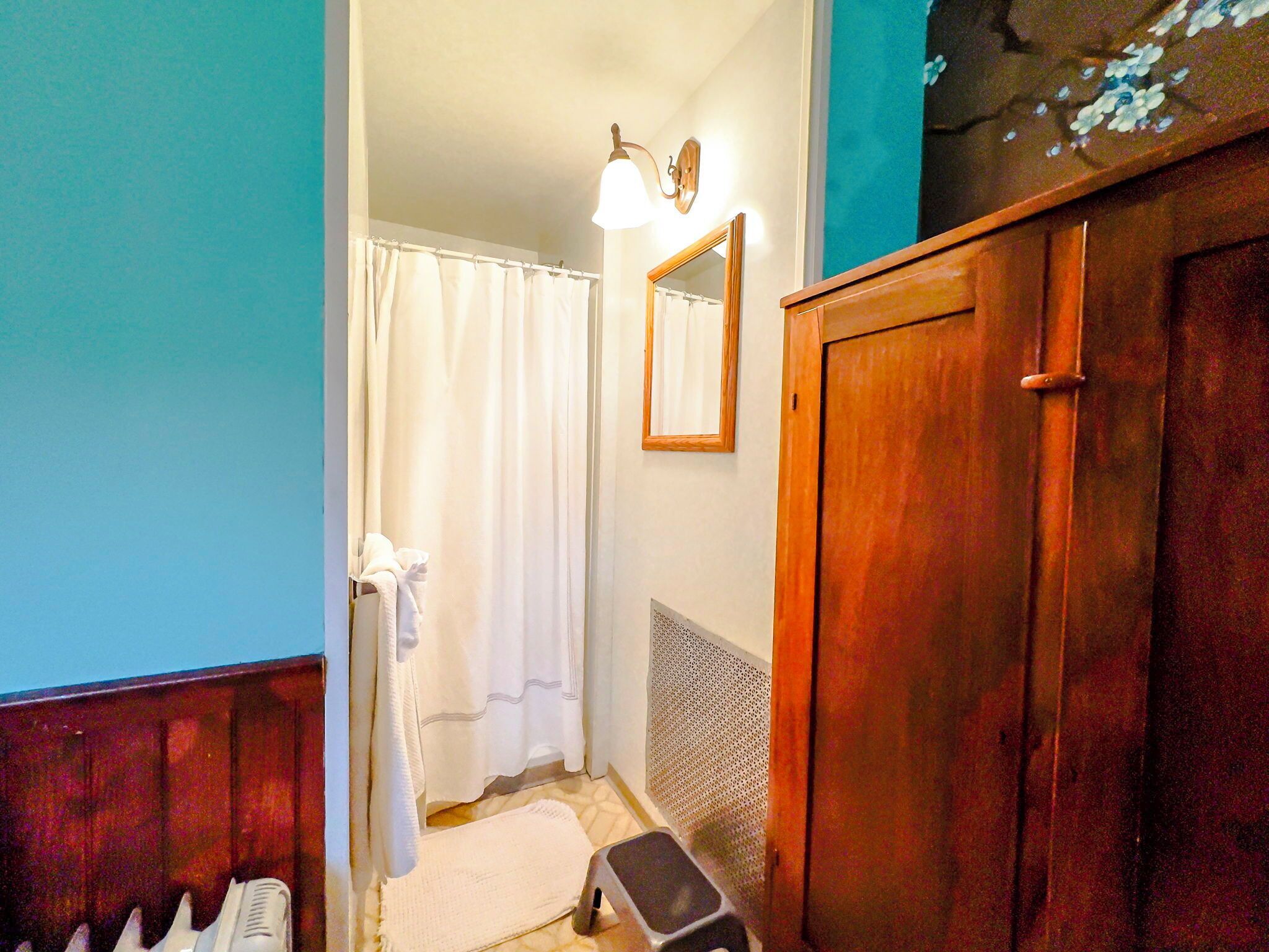A bathroom with a wooden cabinet, a white shower curtain, a small step stool, and a wall-mounted light fixture.