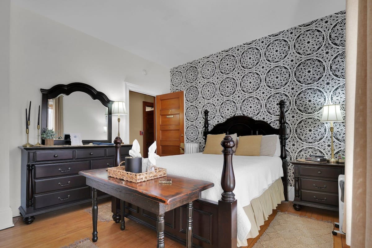 A bedroom with a wooden bed, a dresser, a nightstand, a mirror, and a patterned wallpaper.
