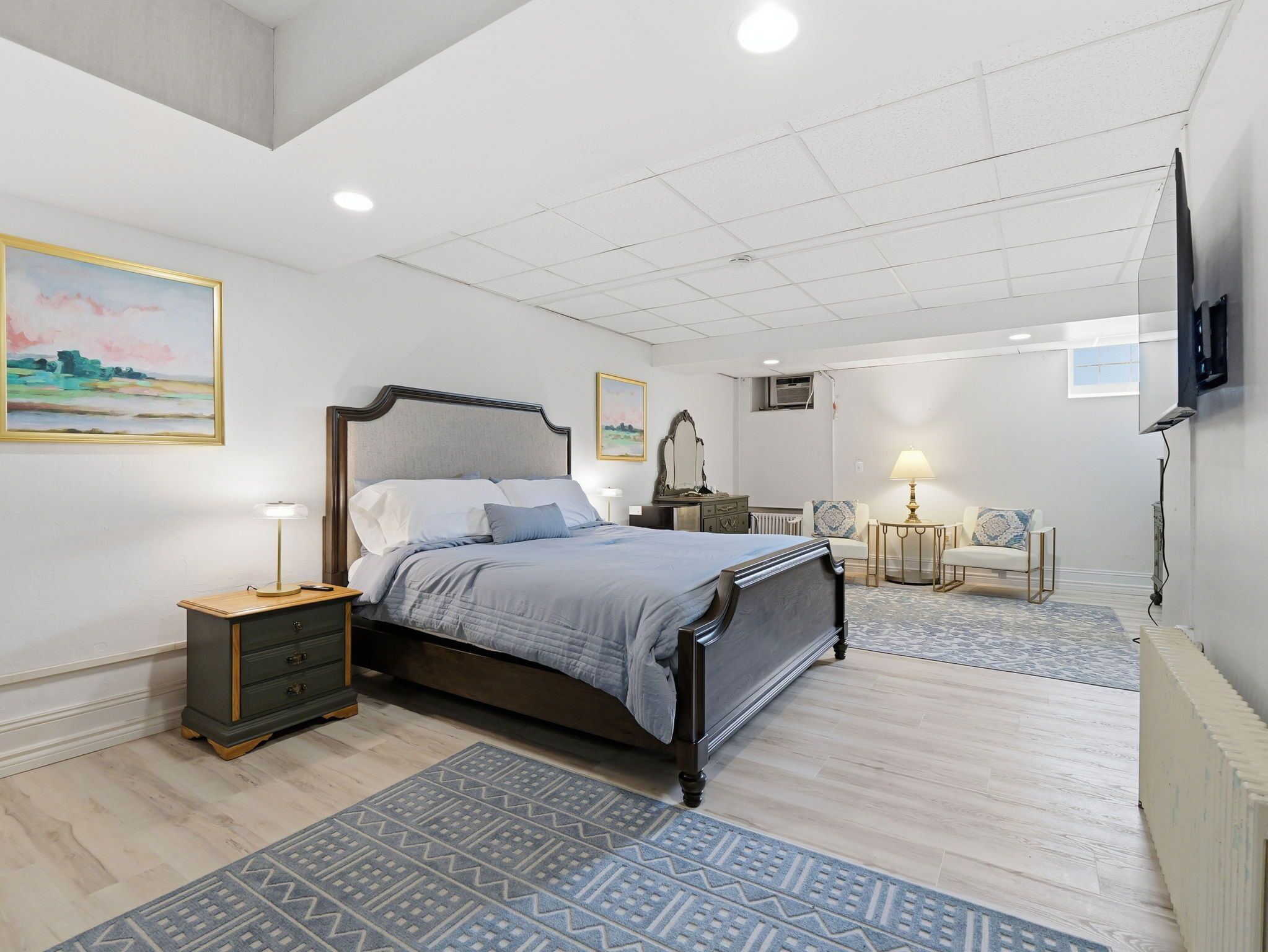 A modern bedroom with a large bed, bedside tables, a dresser, a chair, a TV, and artwork on the walls.