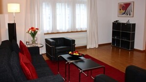 En stue med en sort sofa, røde puder, et sofabord og en skål med frugt. Der er en radiator, en lampe og et billede på væggen.