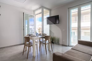 Un salon moderne comprenant un coin repas, un téléviseur à écran plat fixé au mur et de grandes fenêtres offrant une vue sur l’extérieur.