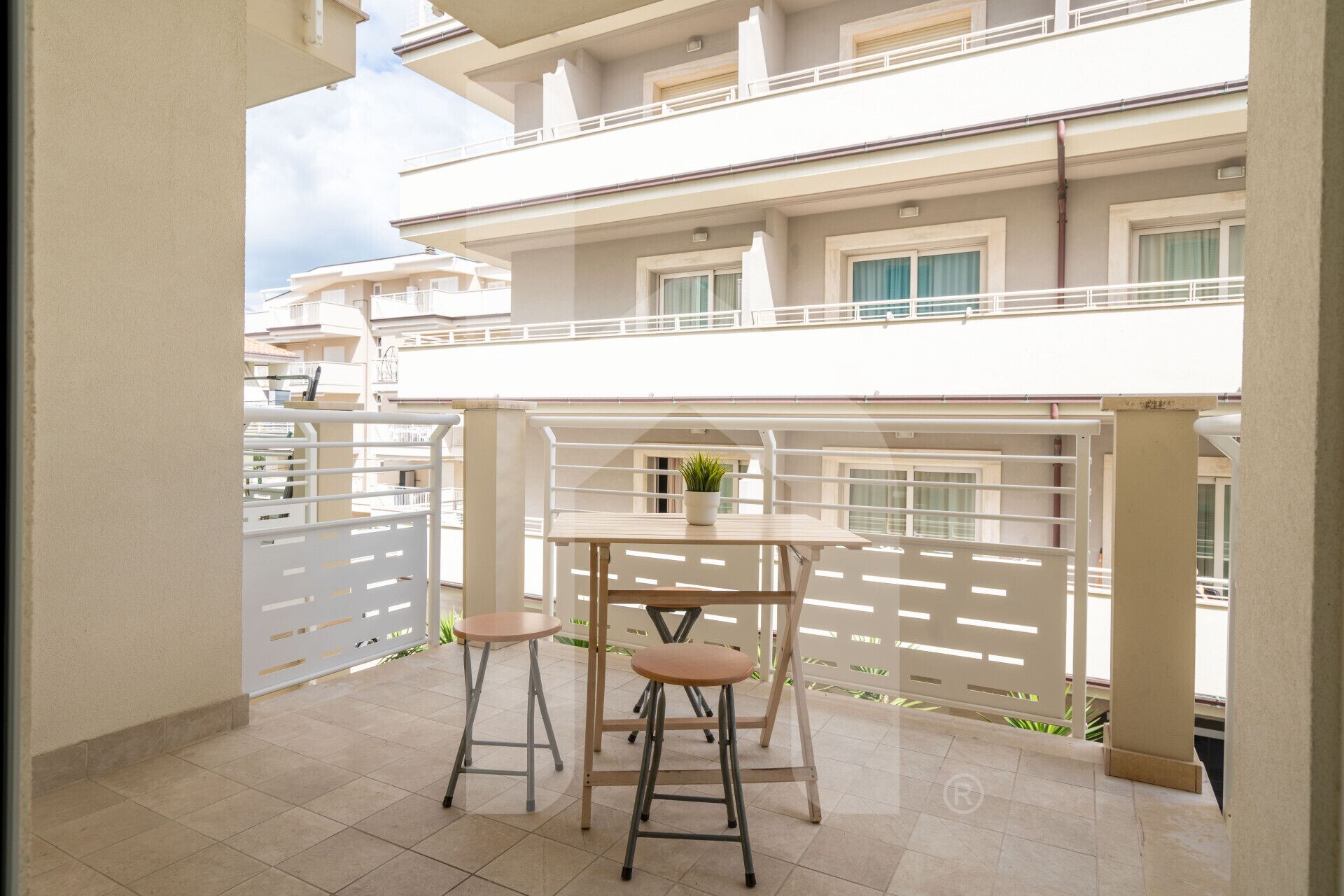 A balcony with a table and two stools, a potted plant, and a view of neighboring buildings.