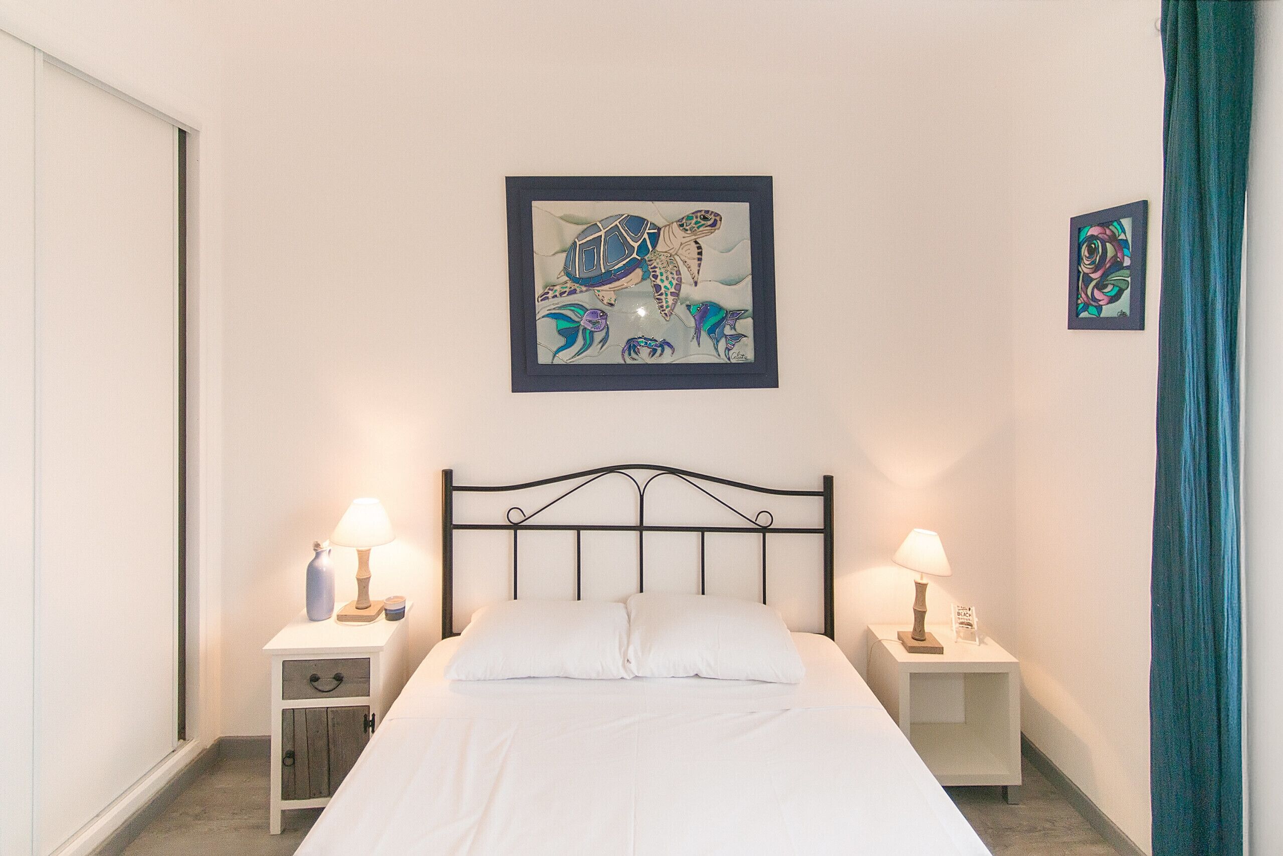Une chambre à coucher avec un lit, deux tables de chevet avec des lampes, un tableau encadré au mur et un rideau.