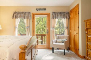 A bedroom with a bed, a chair, a wooden wardrobe, and a door leading to a balcony with a view of trees.