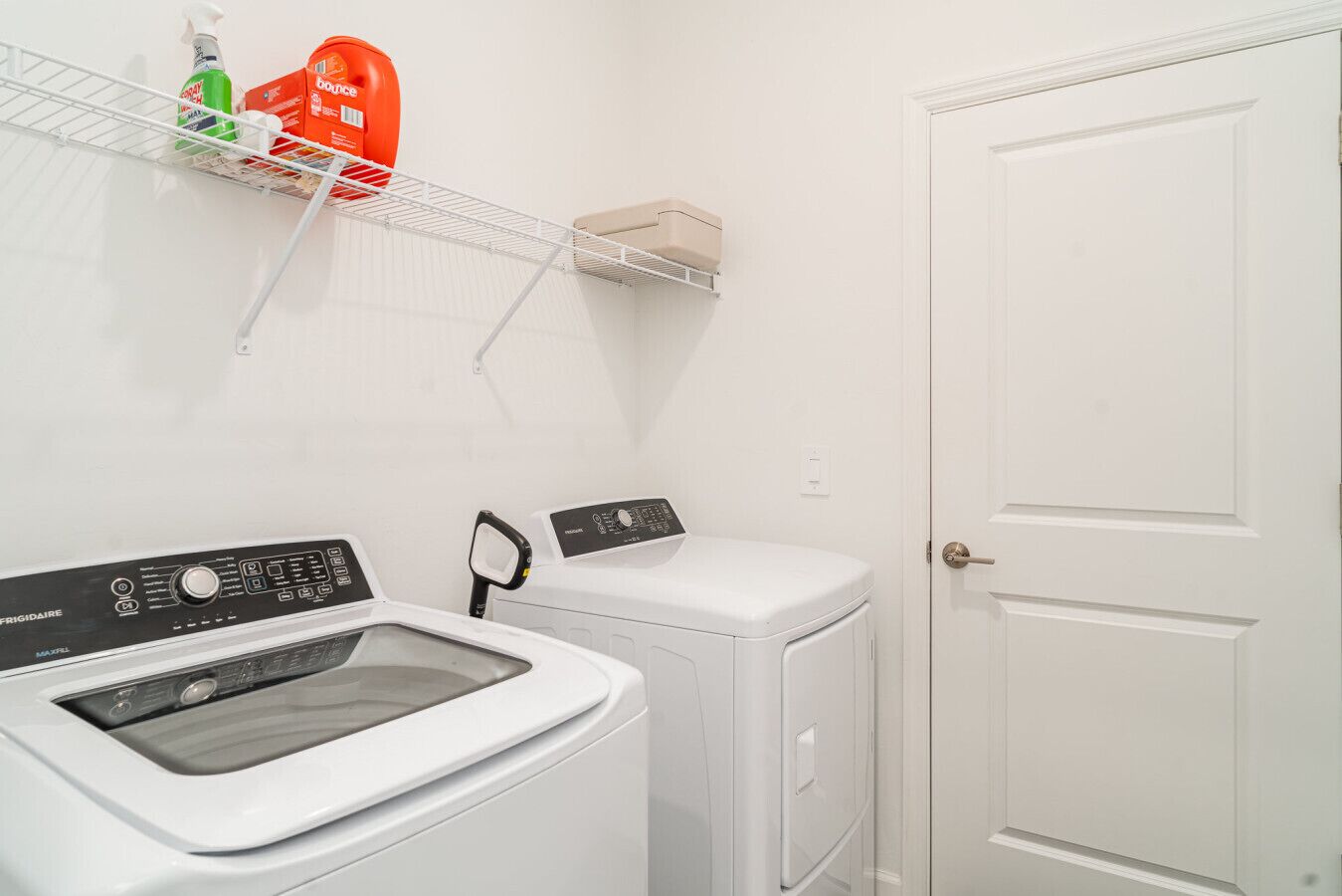 A laundry area with a front-loading washing machine and a dryer, a shelf with cleaning supplies, and a white door.
