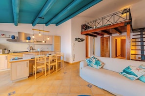 Una cocina moderna con armarios de madera, un comedor con sillas, un sofá con cojines decorativos y un dormitorio en lo alto con balcón.