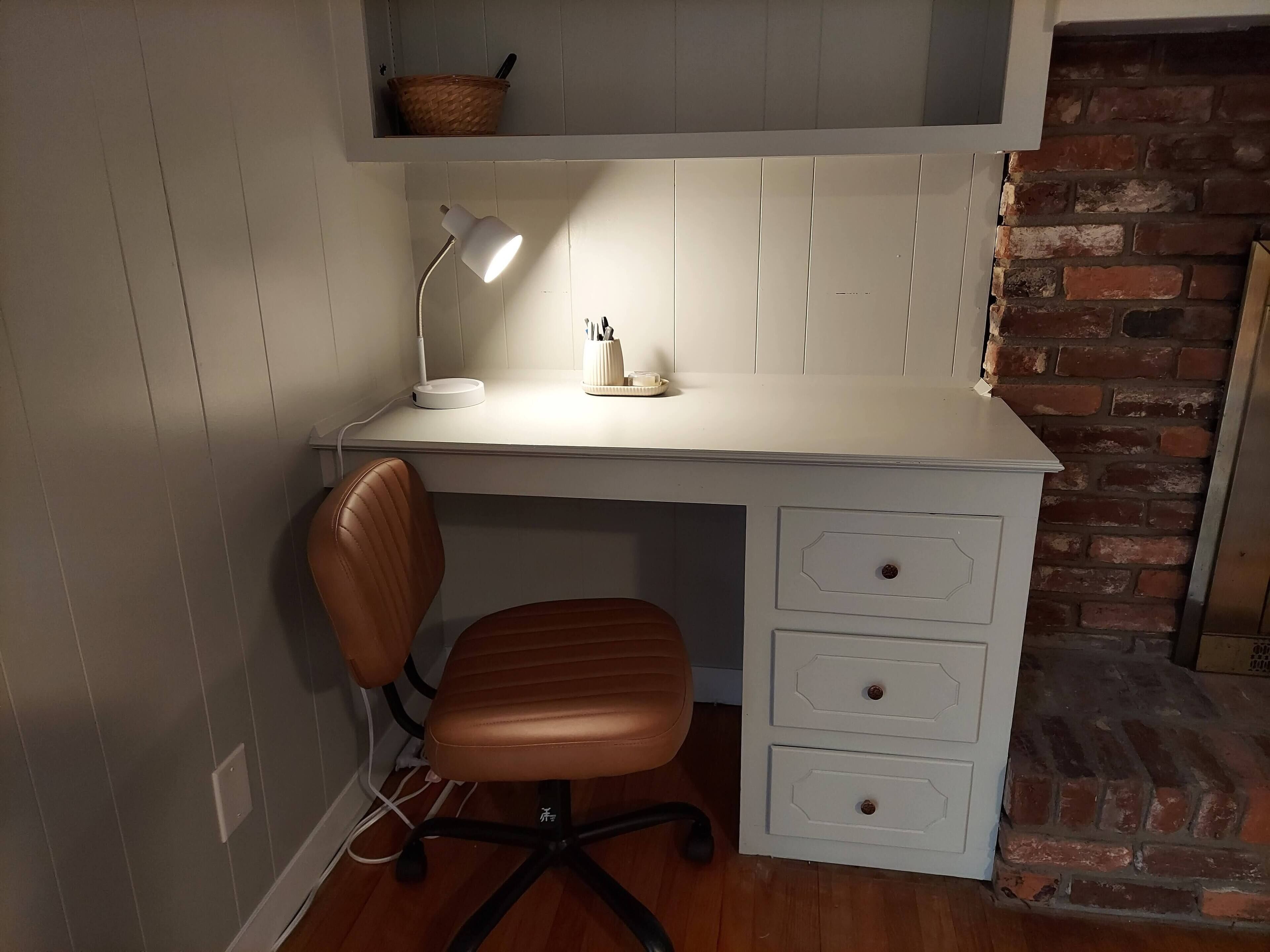A compact workspace with a white desk, a brown leather chair, a desk lamp, and a small basket on a shelf.