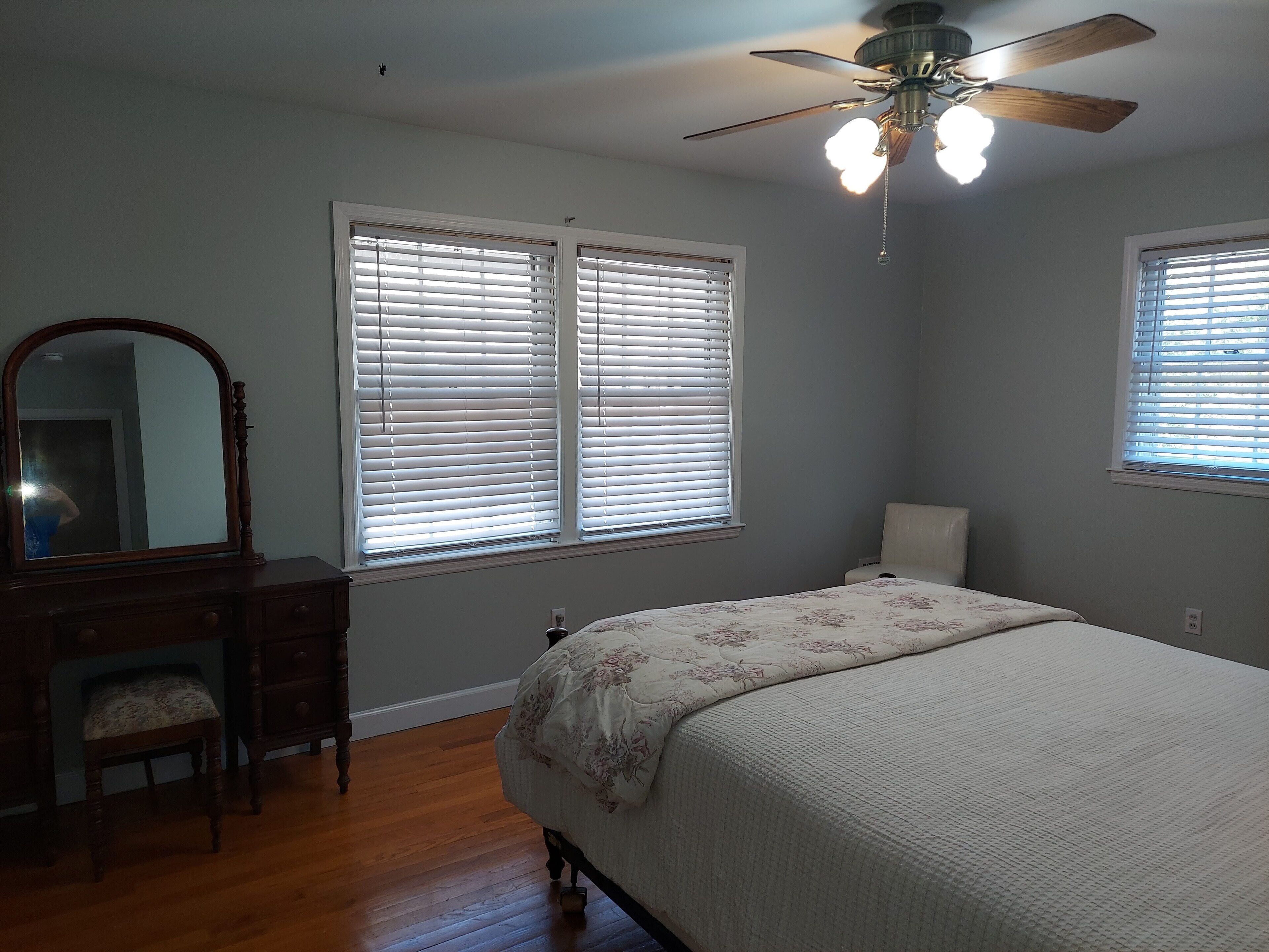 A bedroom with a bed, dresser, mirror, and two windows with blinds.