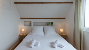Een net opgemaakt bed met witte lakens en twee opgerolde handdoeken, twee bedlampen en een plank met boeken en een lantaarn.