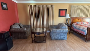 A hotel room with a wooden floor, a sofa, a chair, a bed with a patterned bedspread, a nightstand with a lamp, and a framed picture on the wall. - Vista Motel - Riverside (Riverside)