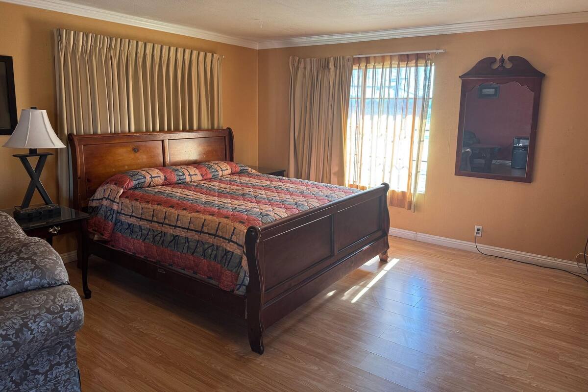 A bedroom with a wooden bed, a nightstand, a lamp, a floral armchair, and a mirror on the wall.