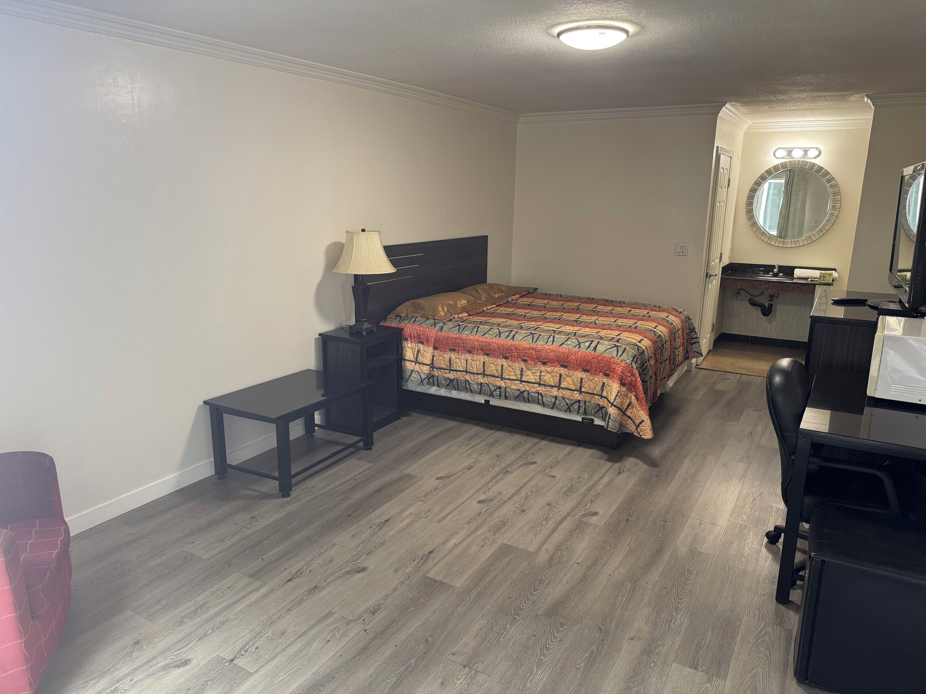 Quarto com cama, mesa de cabeceira, luminária, escrivaninha, cadeira e banheiro com pia e espelho.