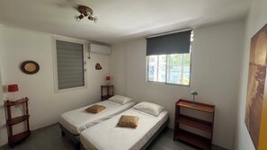 Une chambre avec deux lits, une fenêtre, un ventilateur de plafond, une table de chevet avec une lampe et un climatiseur fixé au mur.