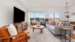 A modern living room with a large flat-screen TV, leather sofas, a coffee table, and a view of the outdoors.