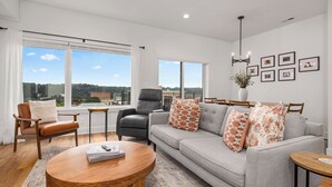 A modern living room with a grey sofa, two armchairs, a coffee table, and a large window offering a city view.