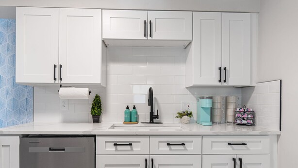 Cozinha moderna com armários brancos, lava-louças em aço inoxidável, torneira preta e painel de azulejos brancos.