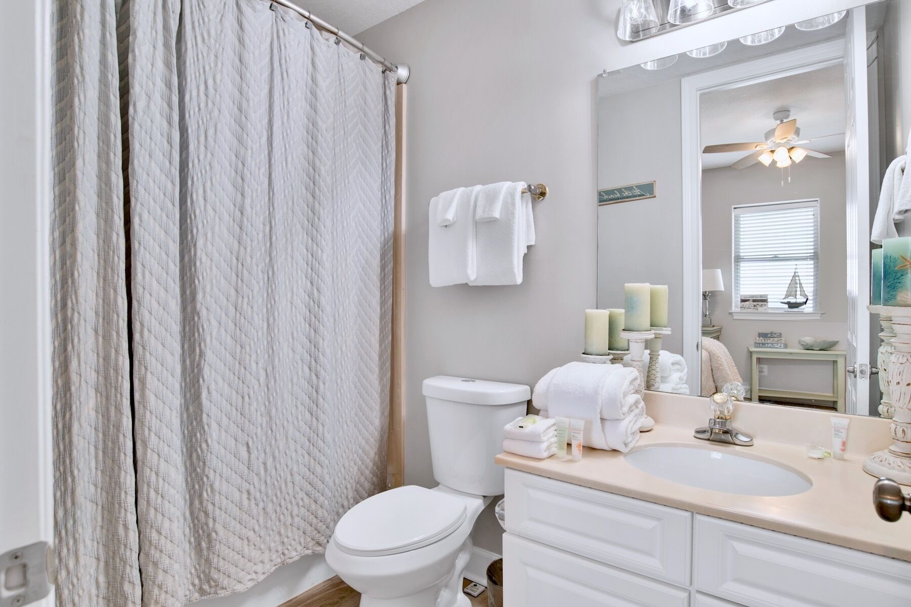 A bathroom with a white toilet, a sink with toiletries, and a mirror reflecting a ceiling fan and window blinds.