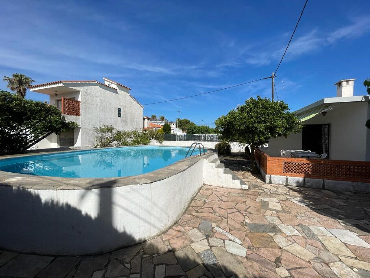 Una piscina con solarium in pietra, circondata da un edificio bianco con tetto in tegole rosse, e un cielo azzurro limpido.