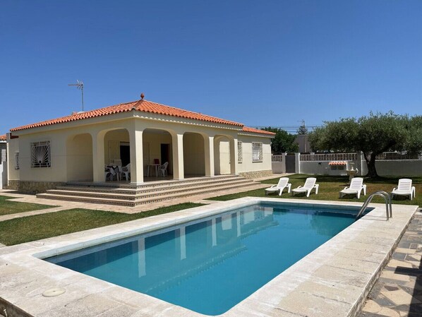 A poolside area with a pool, a covered patio with chairs, and a building with a tiled roof.