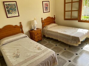 A bedroom with two single beds, a nightstand with a lamp, and a window with a view of greenery.