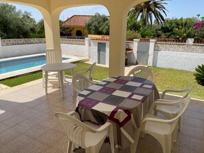 A covered patio area with a pool, outdoor seating, and a view of a house with a red-tiled roof.