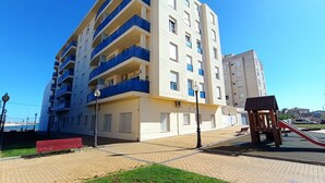 Un edificio residencial de varios pisos con balcones, un parque infantil y un banco rojo.