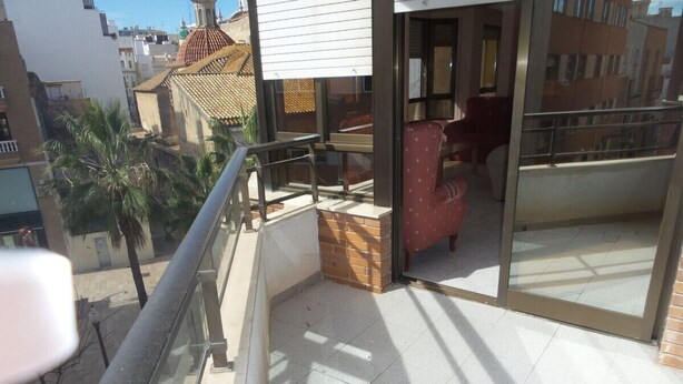 Ein Balkon mit Glastür zu einem Innenraum, ein roter Stuhl und Blick auf benachbarte Gebäude und Palmen.