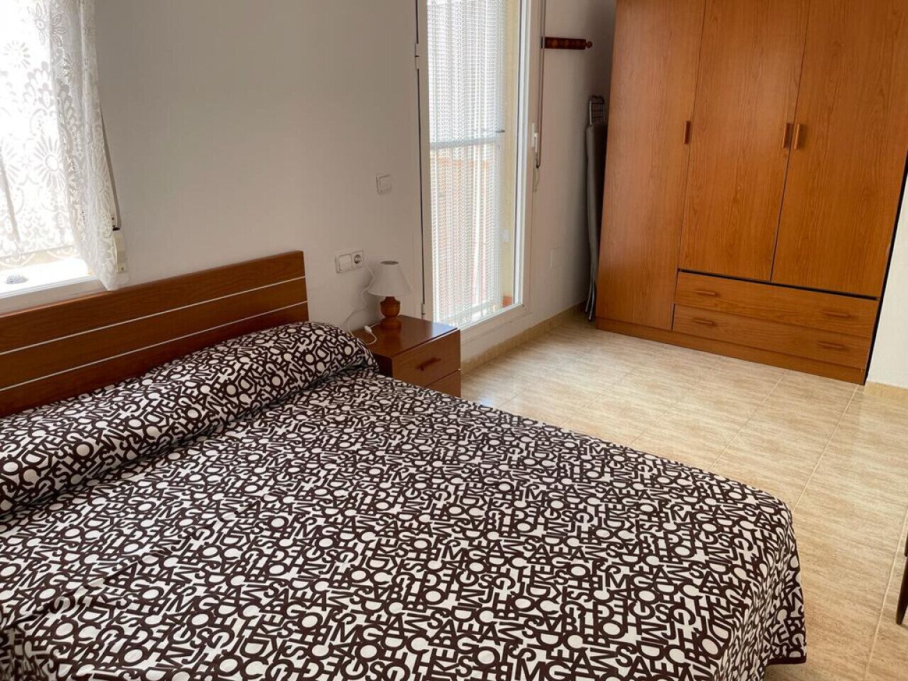 Een slaapkamer met een bed, een nachtkastje, een houten garderobe en een raam met gordijnen.