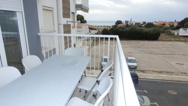 Un balcon avec une table et des chaises blanches, donnant sur un parking et une plage.
