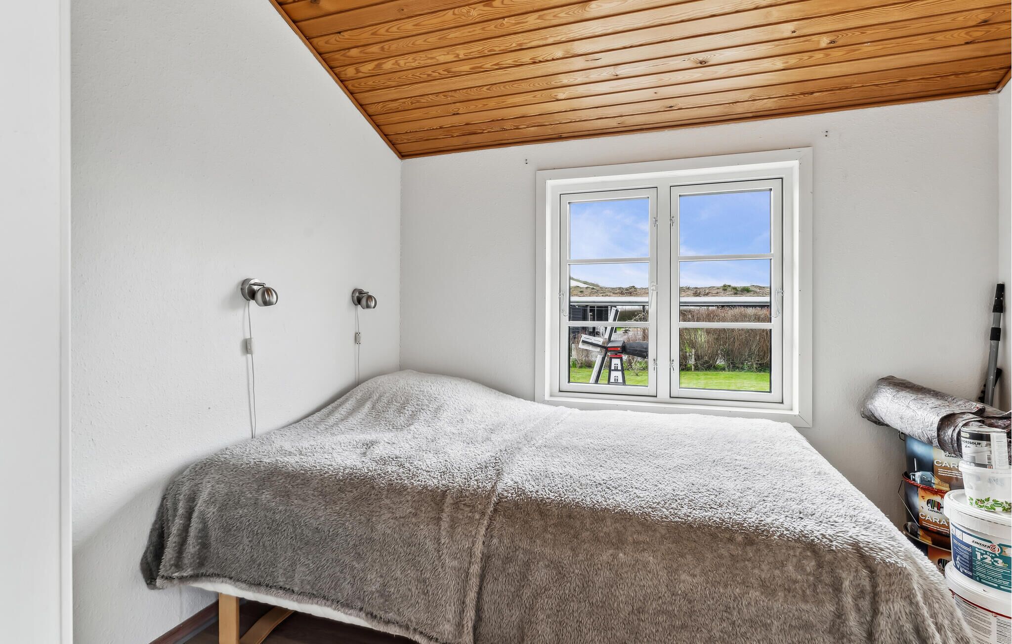 A bedroom with a wooden ceiling, a large window with a view of a house and landscape, and a bed with a grey blanket.