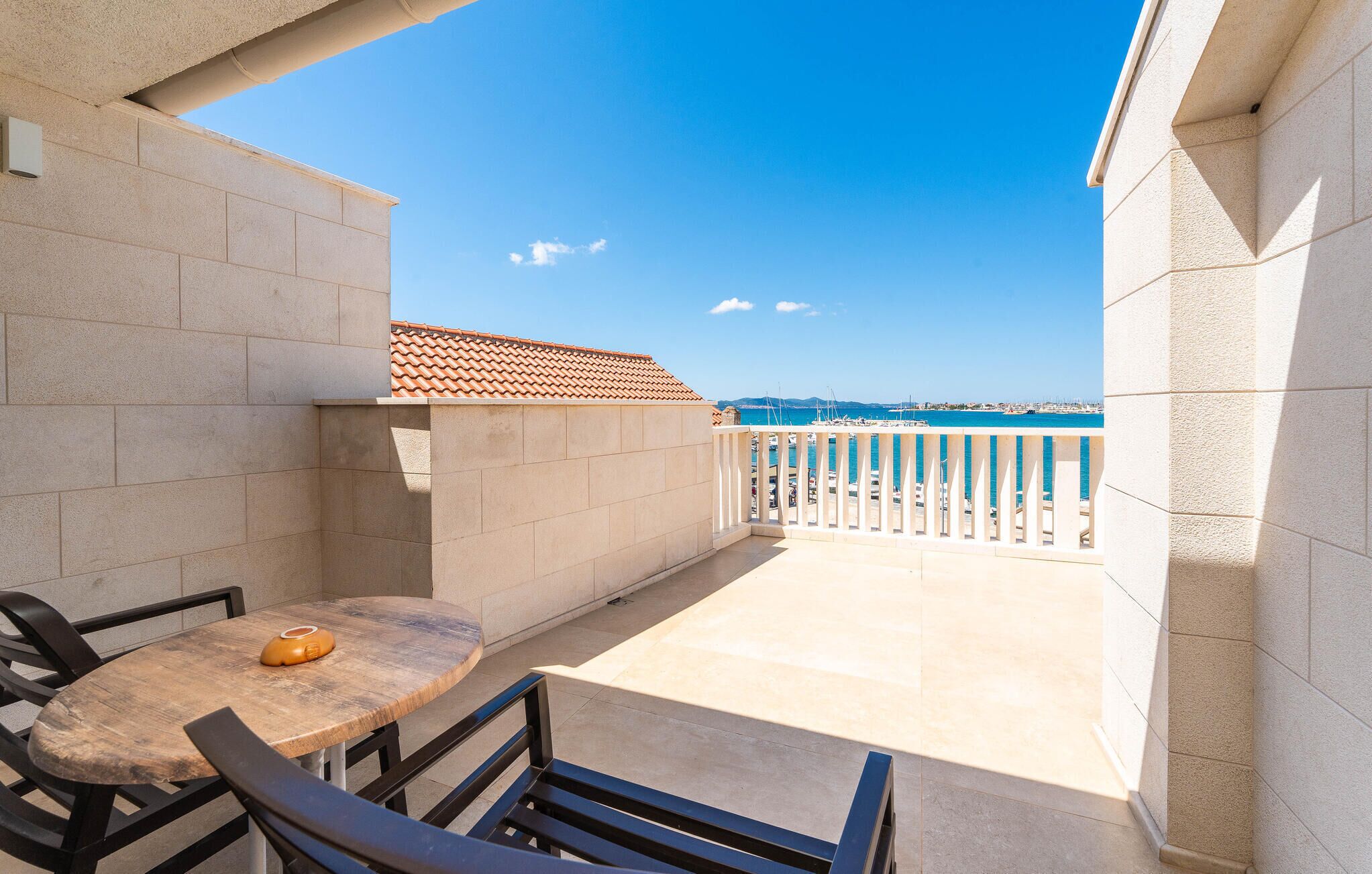 A balcony with a view of the sea, a table with a chair, and a tiled floor.