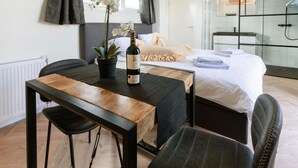 A modern hotel room with a wooden dining table, two chairs, a potted orchid, and a bottle of wine. The room features a large bed with a white comforter, a glass shower enclosure, and a view of the city. - Cozy home in Vinkeveen with sauna (Vinkeveen)